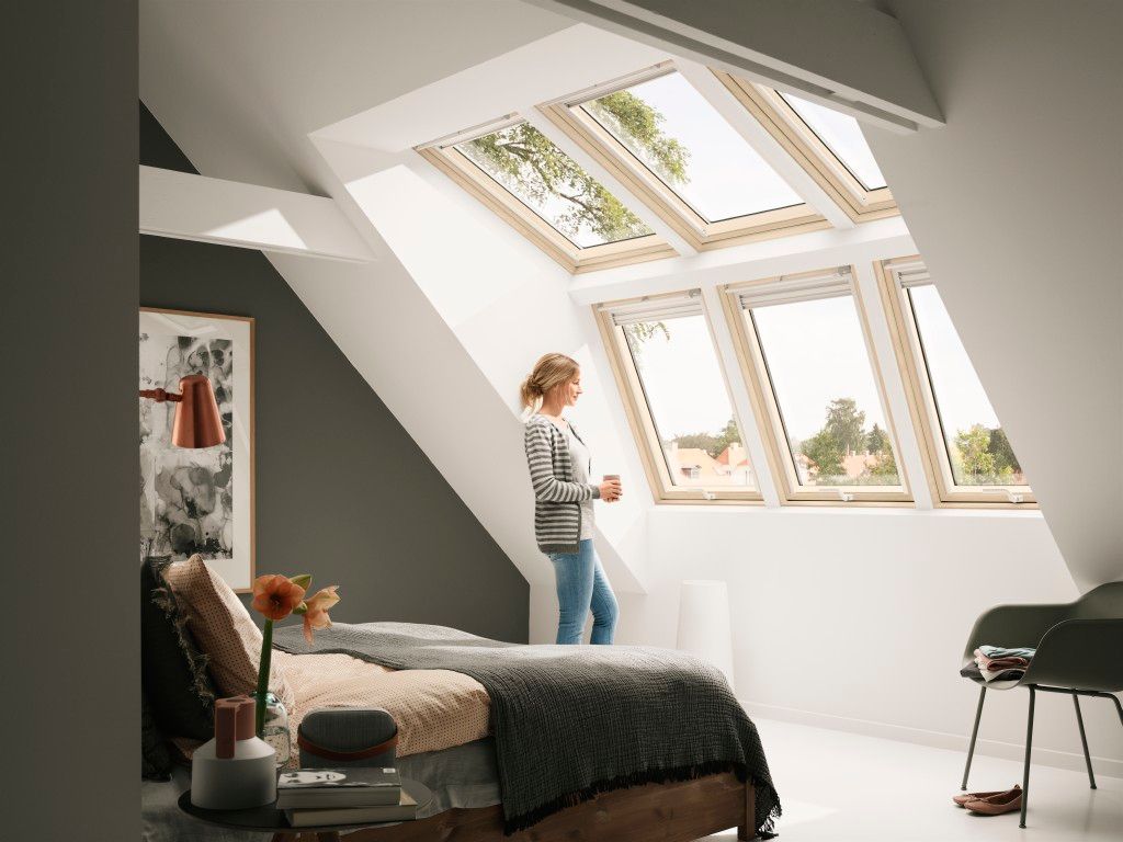 Eine Frau steht im Schlafzimmer vor einer Front aus sechs Dachfenstern, die in drei Reihen zu je zwei Fenstern übereinander angeordnet sind – ein lichtdurchfluteter Raum mit modernem Design.