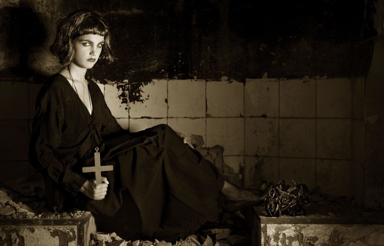 Thierry Aguiar : Photo sépia d'Albane en gothique, assise de profil dans une cheminée, en robe noire,  un cruciifix en bois dans la main droite, un bouquet de roses noires déposées à ses pieds.