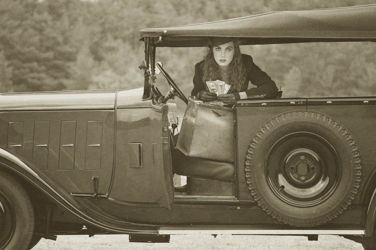 Thierry Aguiar : Photo noir et blanc délavé d'Albane en Bonnie, dans une Citroën C4 Torpedo conduite à droite de profil, à genoux de face sur le siège, comptant des dollars dans un sac en cuir, portant un chapeau de Lilô Chapô à Grasse et des mitaines.