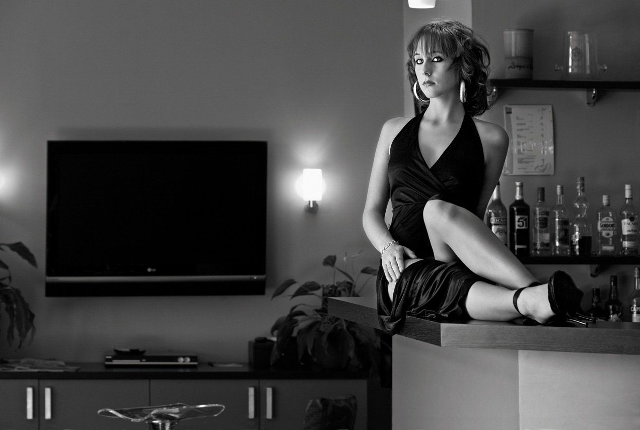 Photo noir et blanc d'Audrey en robe noire fendue, portant des chaussures à talons aiguilles et des boucles d’oreilles créoles, assise de face sur le bar de l’hôtel Mandarina à Grasse.
