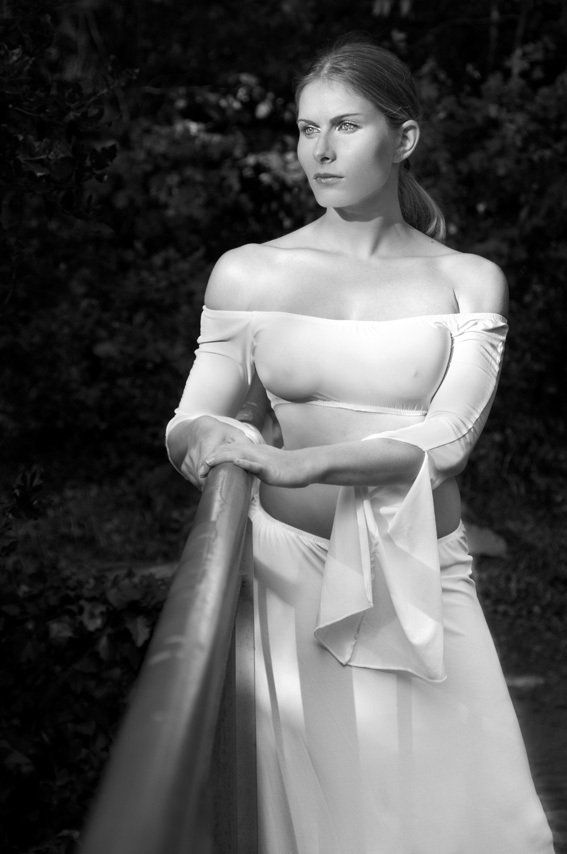 Aguiar Thierry : photo d'Aurélie en robe blanche sexy, debout de face sur un pont en métal, appuyée sur la balustrade, regardant au loin