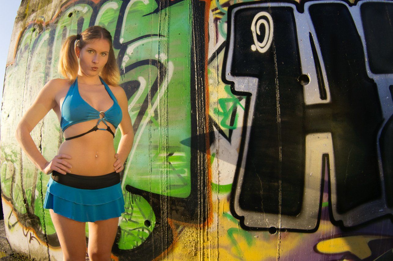 Thierry Aguiar. Aurélie avec des couettes debout au soleil couchant devant un mur tagué, portant un bustier et une robe bleus