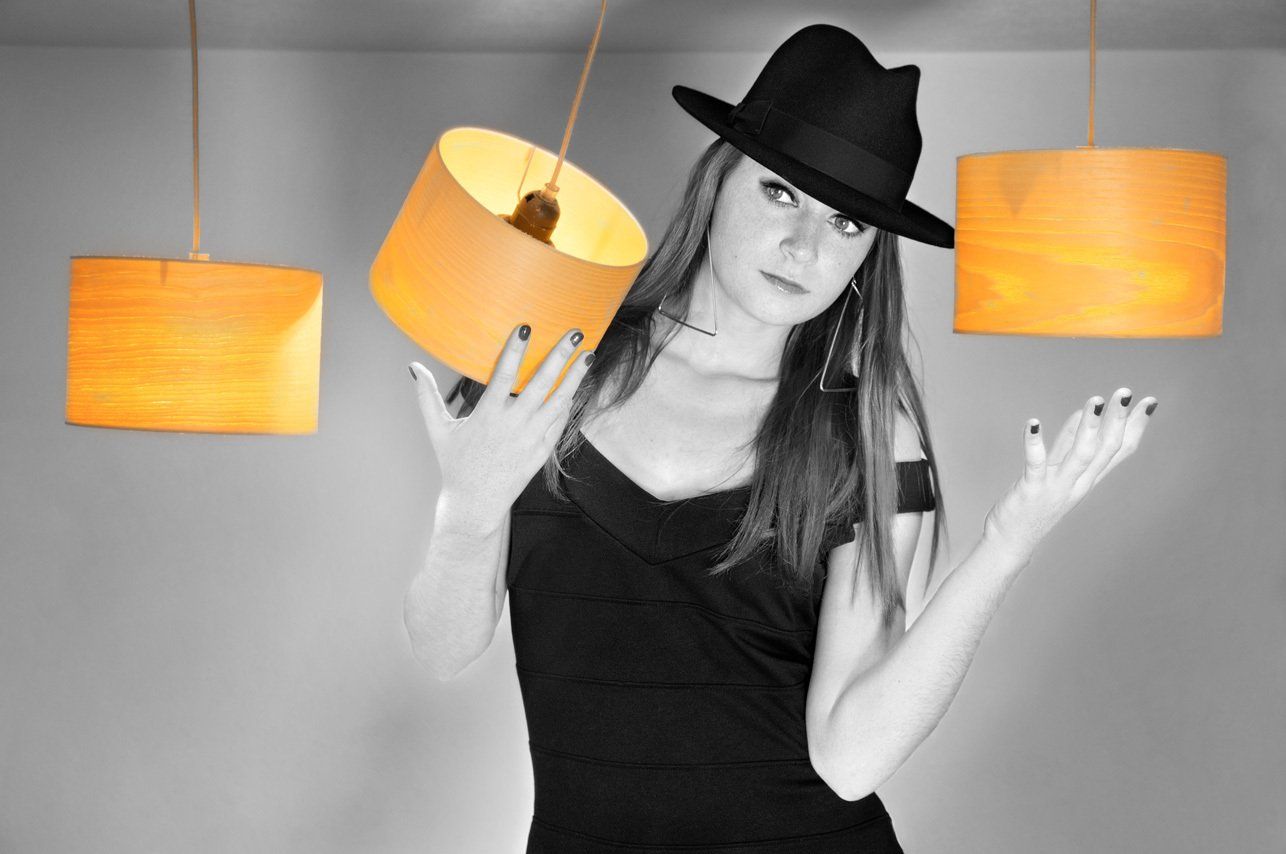 Axelle portant un chapeau et une robe noirs, des boucles d'oreilles en triangle, encadrée par des lumières suspendues.