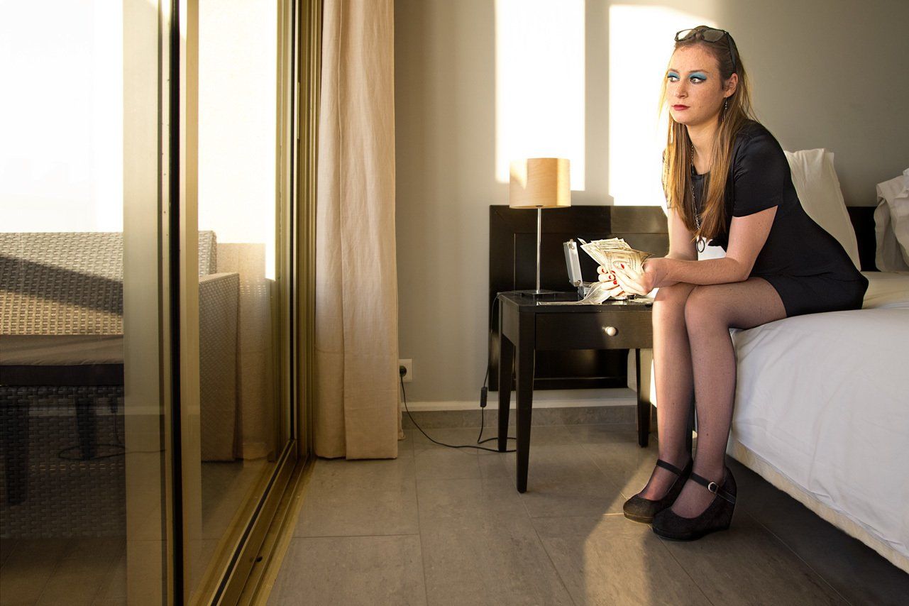 Thierry Aguiar : Photo couleur d'Axelle portant une robe noire, assise sur le lit côté, d’une chambre d'hôtel, des dollars dans les mains, regardant dehors, des lunettes de soleil dans les cheveux..