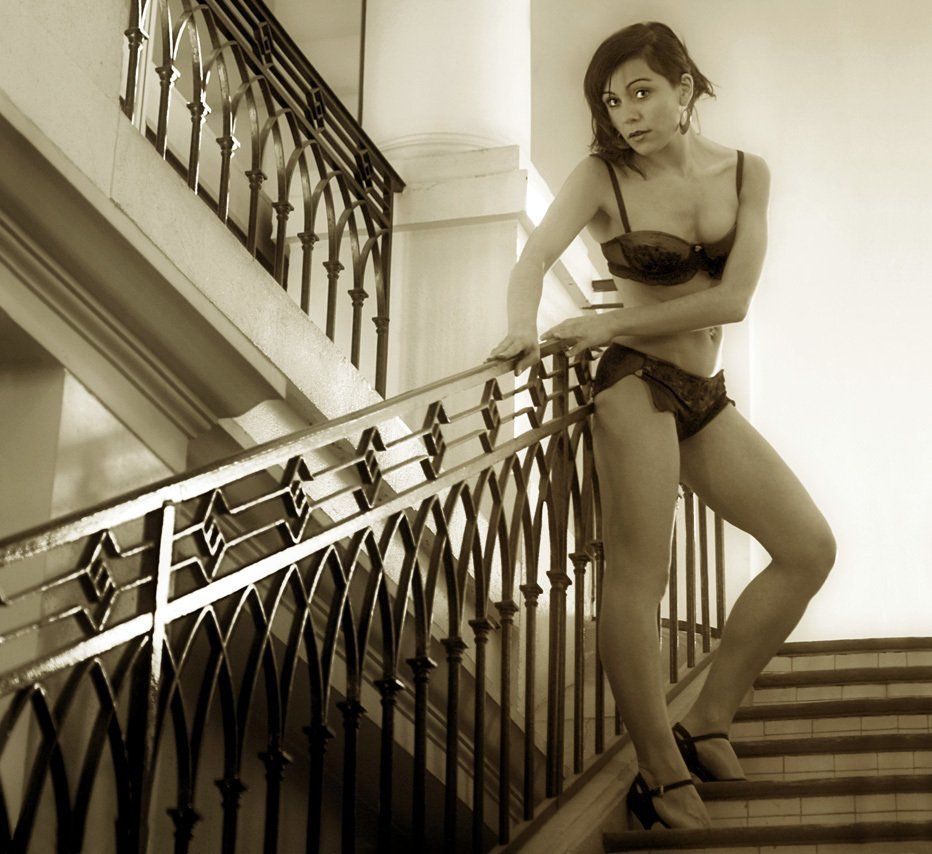 Photo sépia de Céline debout en sous-vêtements noirs, se tenant à la rampe de l’escalier dans un bâtiment du 18ème siècle.