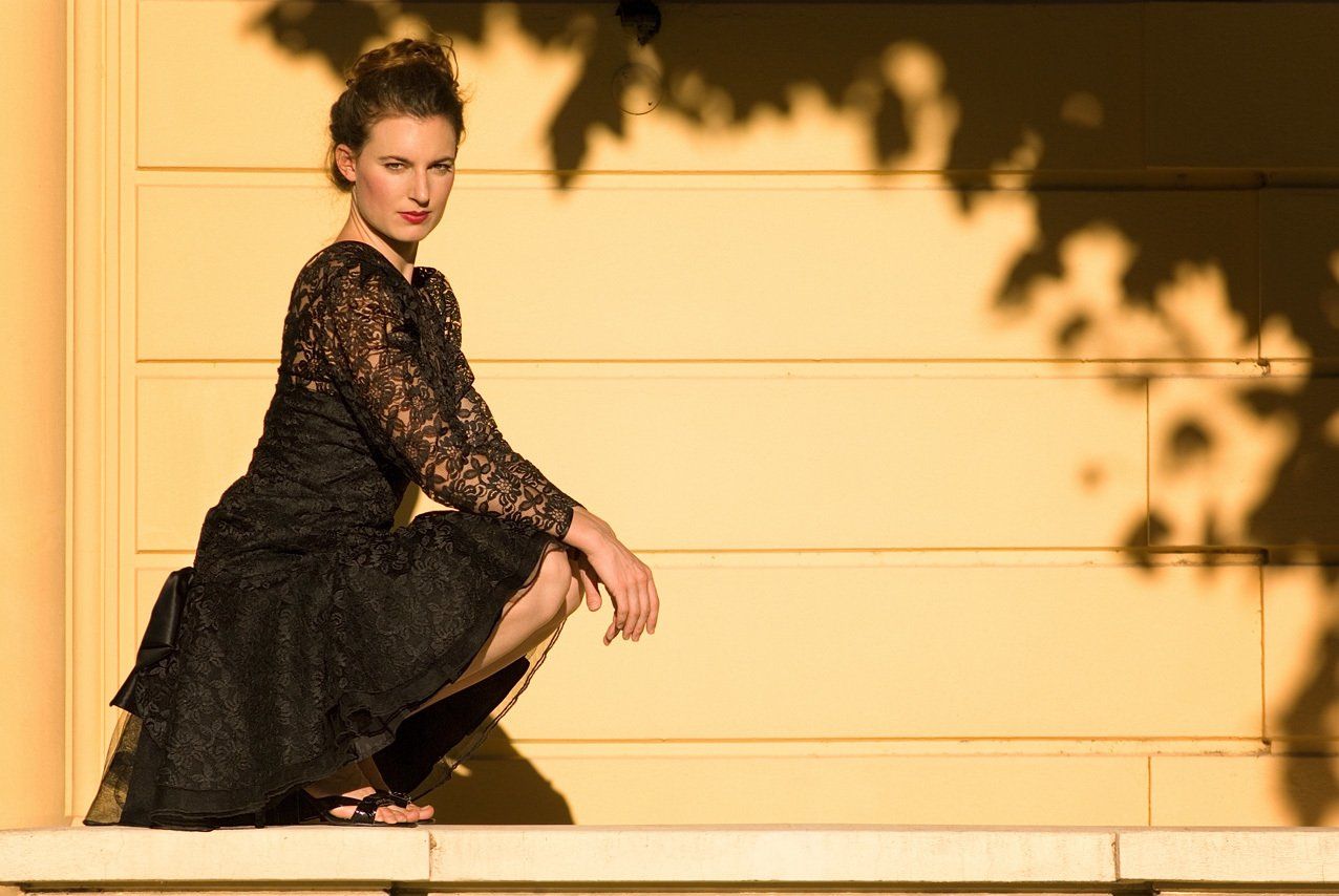 Thierry Aguiar : photo de Charlotte accroupie de profil sur un muret, devant un mur en pierre jaune, ausoleil couchant, portant une robe noire à dentelle. Photo du jour sur ITIS.