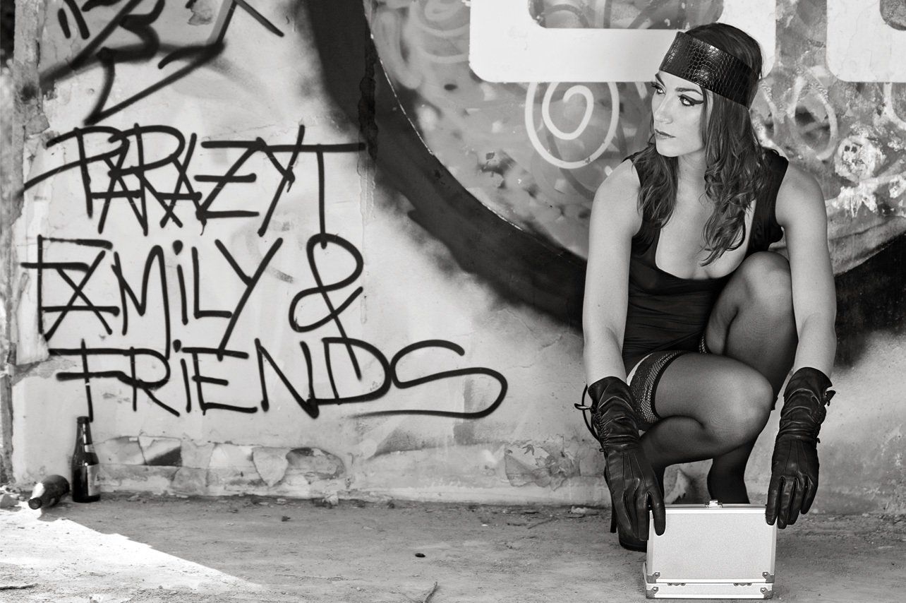 Claudia à genoux de face devant un mur taggué, regardant à sa droite, ouvrant une mallette en aluminium un pistolet à la main