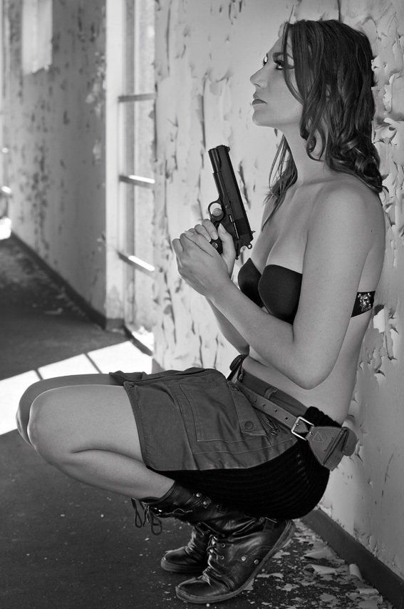 Thierry Aguiar : Photo noir et blanc de Claudia accroupie adossée à un mur, regardant devant elle, tenant un pistolet à deux mains, habillée d'un soutien-gorge noir et d'une jupe noire et marron.