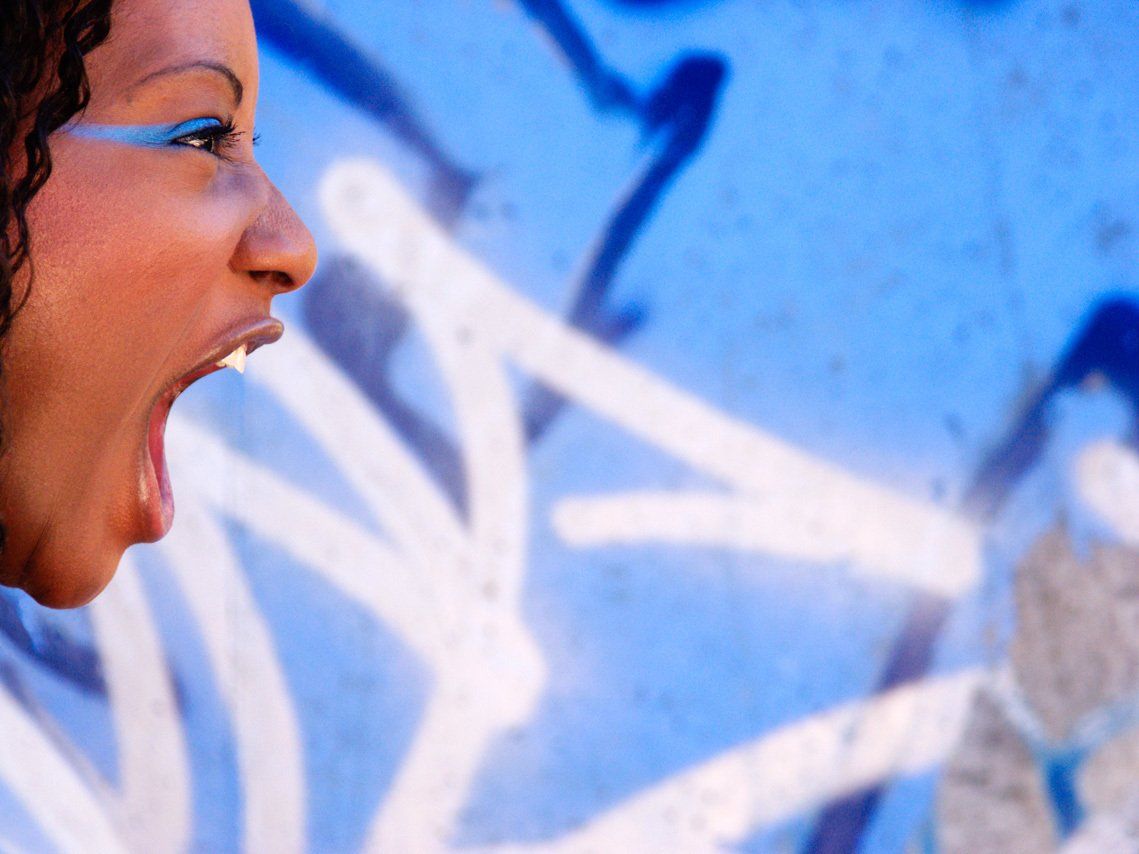 Thierry Aguiar : Portrait de profil en lumière naturelle de Jessyca, jeuen fille noire d'origine congolaise, en extérieur, en train de crier devant un mur bleu taggué.