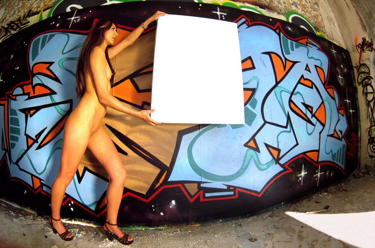 Thierry Aguiar : photo de Juliana nue, debout de profil devant un mur tagué de la maison de retraites abandonnée Riviera à Saint Cézaire sur Siagne, tenant une toile blanche en l’air.