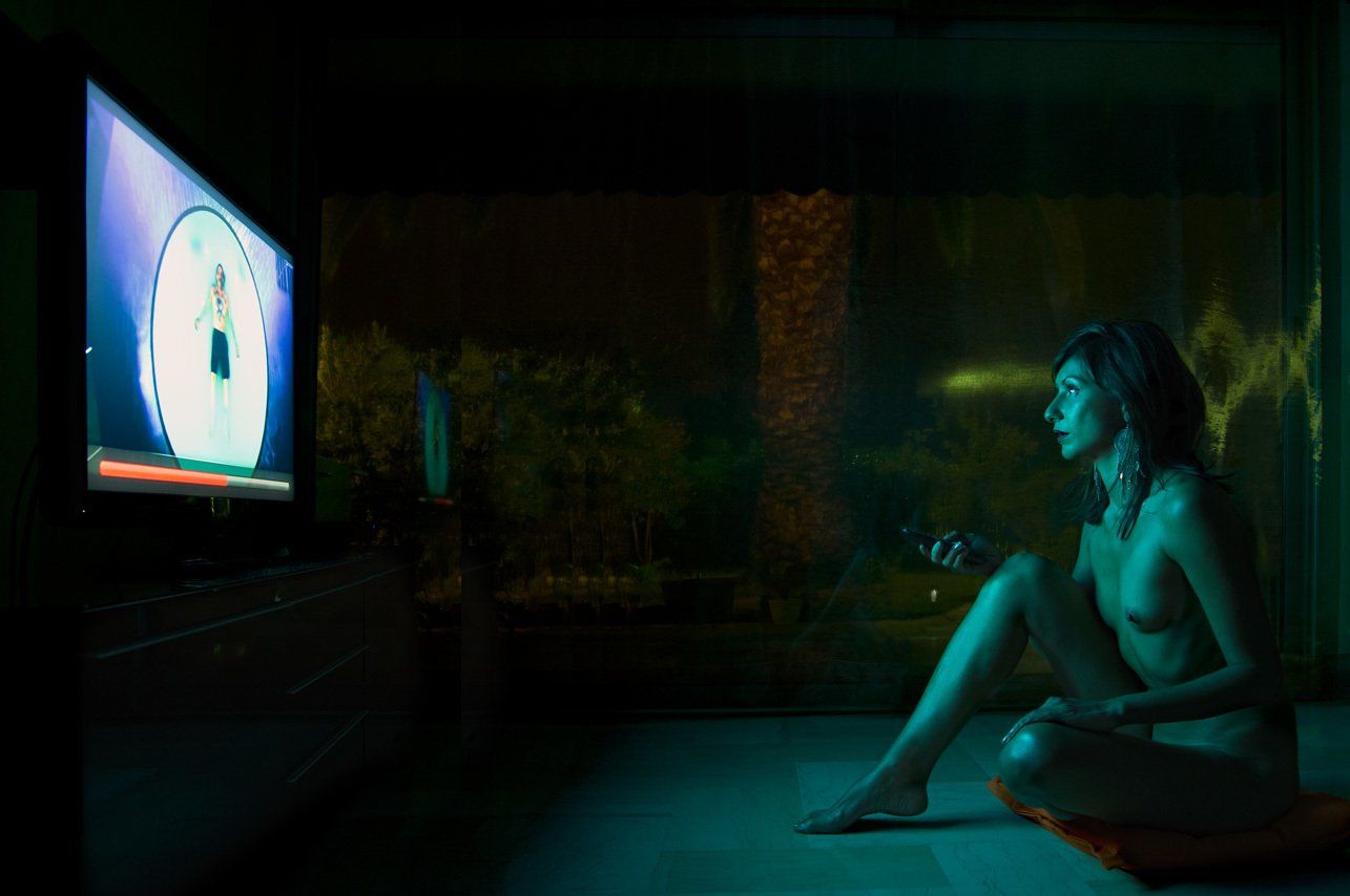 Thierry Aguiar : photo de nuit de Juliana  de profil, nue, assise parterre en train de regarder la télé, la télécommande à la main.