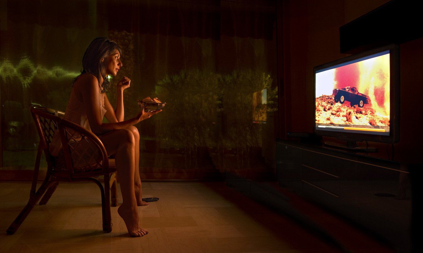 Photo  de Juliana portant une nuisette de soie, assise sur un fauteuil en osier, mangeant des gâteaux en regardant la télé.