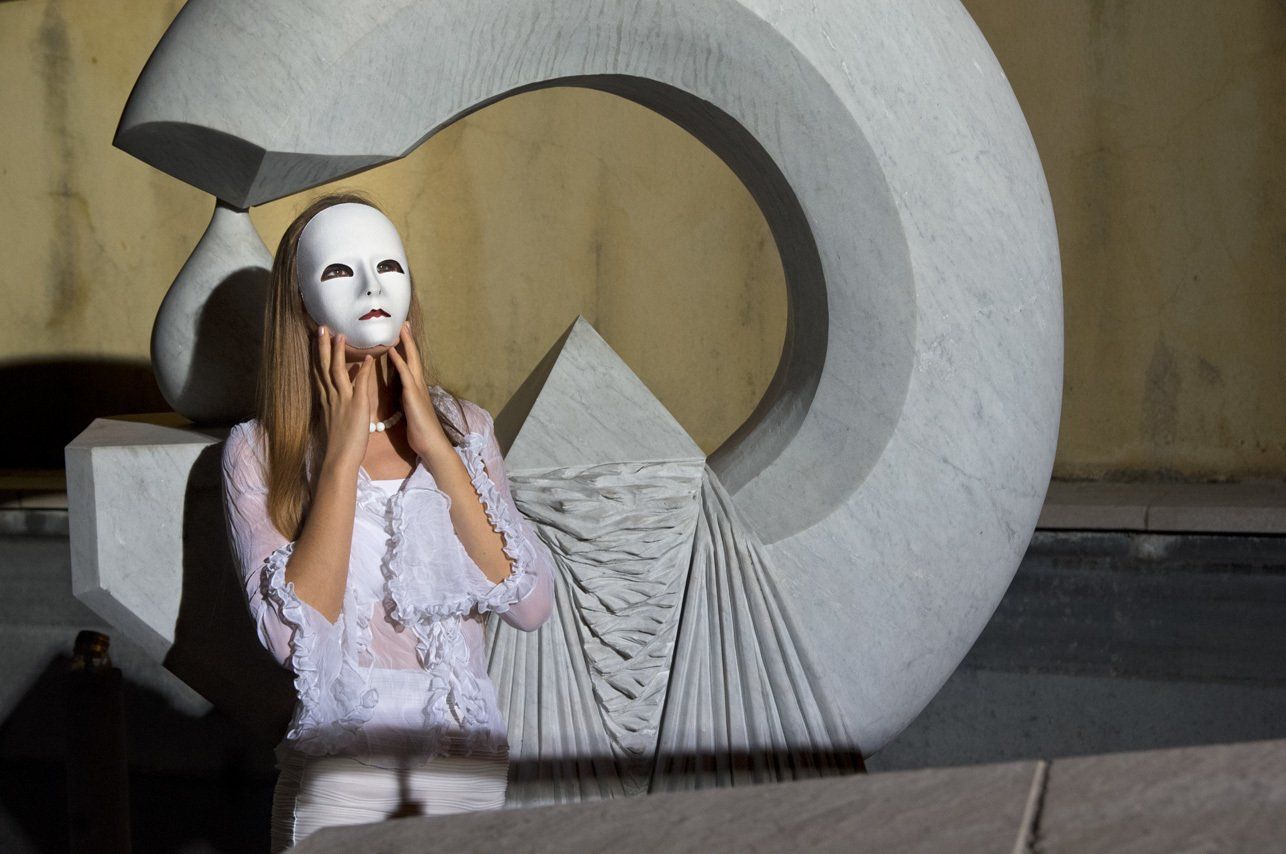 Thierry Aguiar : Photo couleur en plan américain de Kasandra de nuit,  debout devant une sculpture en marbre de bernard verhaeghe, tenat un masque blanc devant son visage, regardant en l'air.