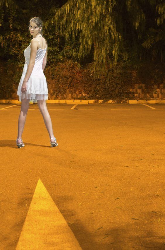 Thierry Aguiar : Photo couleur de plain-pied de Kasandra de dos se retournant, la nuit, sur un parking, portant un bustier, un short, et un chemisier blanc.