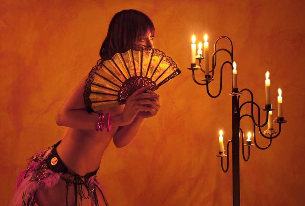 Thierry Aguiar : photo de Laëtitia de profil, nue, debout devant un chandeler, portant une jupe en plumes, un éventail masquant le bas de son visage, éclairée par des bougies.