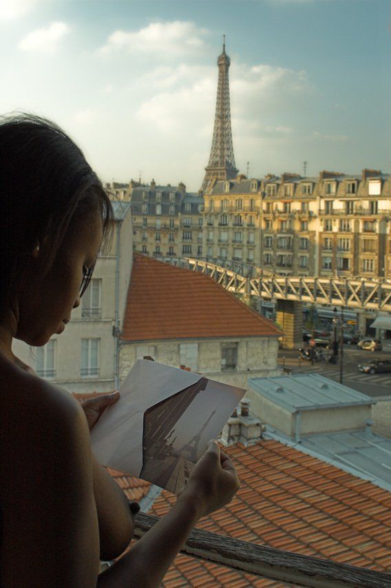 Thierry Aguiar : photo de Lilou, jeune fille noire, nue devant uen fenêtre d'un hôtel dans le 15ème arrondissement de Paris, une carte postale représentant la tour Eiffel à la main, le métro aérien en fond.
