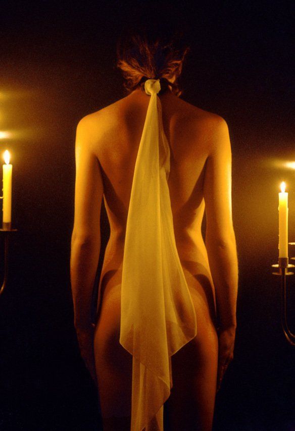 Thierry Aguiar : photo de Maëlle debout nue de dos,  un foulard blanc attaché dans les cheveux descendant sur ses fesses, éclairée par des bougies,  deux chandeliers à ses côtés.