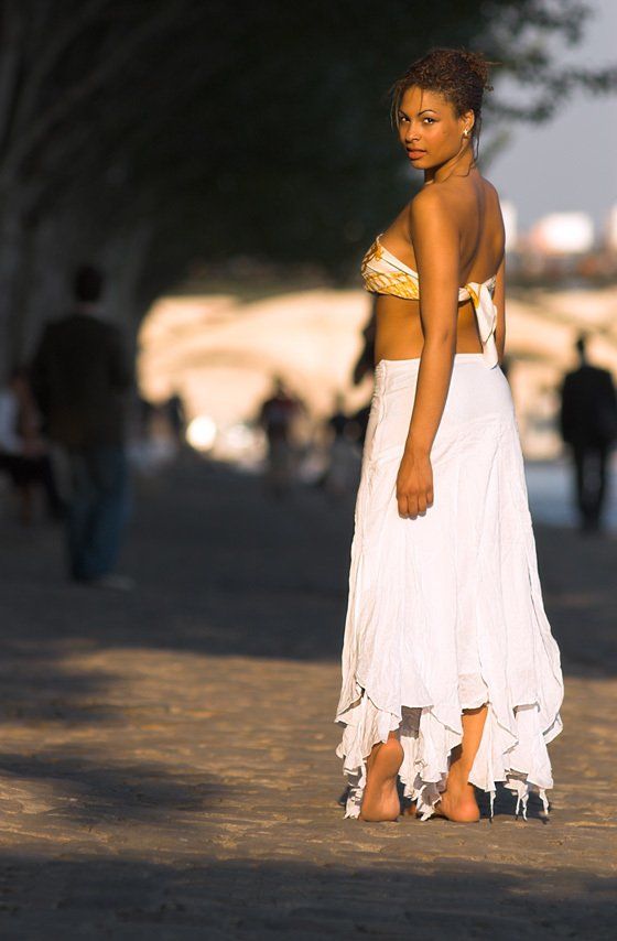 Thierry Aguiar : Photo de Magali au soleil couchant, jeune fille noire, portant un bustier coloré et une robe blanche,  marchant pieds nus le long de la Seine, de dos, se retournant, le pont des arts en arrière-plan.