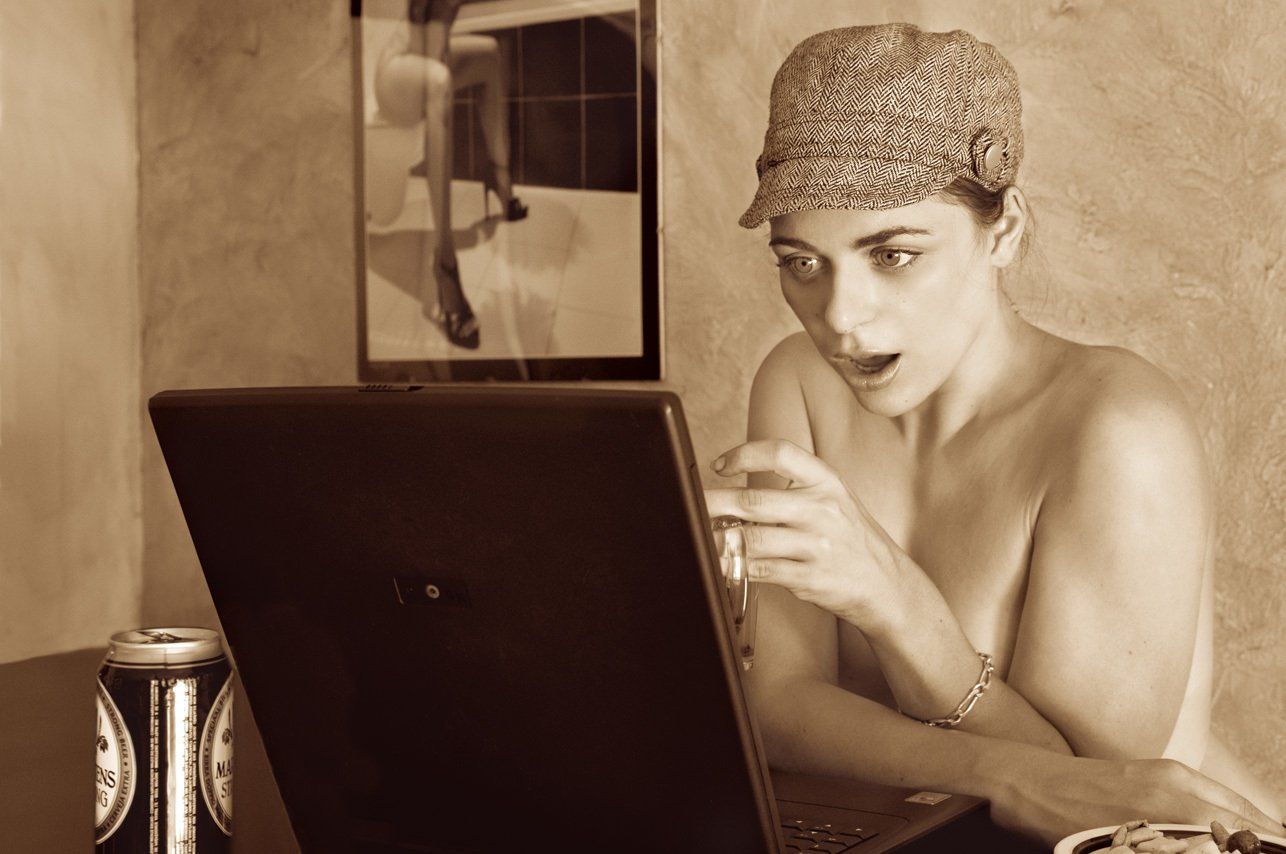 Thierry Aguiar : photo sépia de Manéa, jeune fille aux yeux verts, assise nue à un bar, portant une casquette en tweed, regardant son ordinateur une chope à la main, une canette posée à côté l’ordi.
