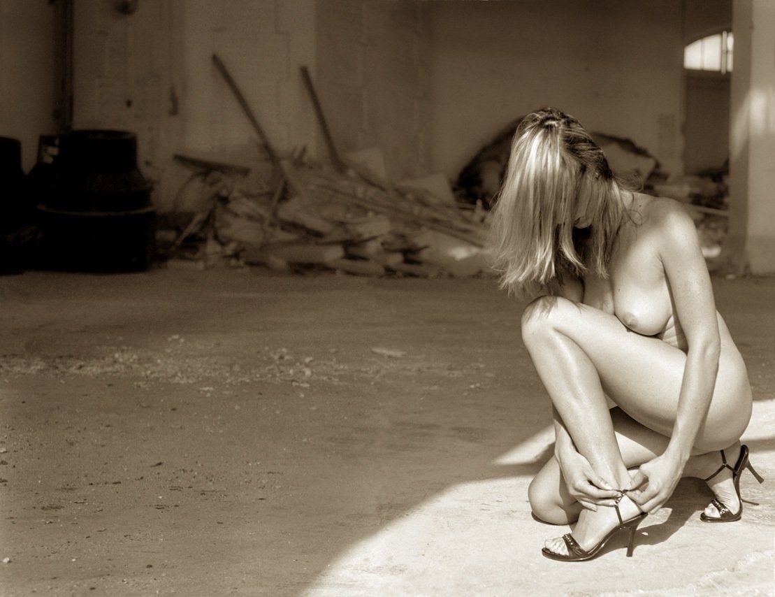 Thierry Aguiar : photo sépia de Sabrina nue, accroupie de profil en train d’attacher sa chaussure, dans un bâtiment de Roure à Grasse.