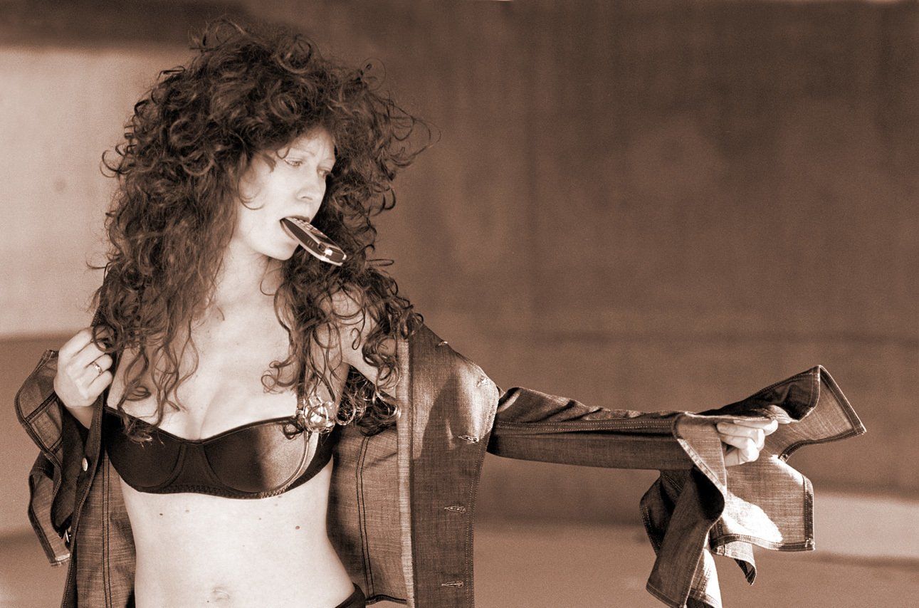 Valérie de profil, en soutien-gorge dans un bâtiment abandonné,  enfilant une veste en jean, son téléphone dans la bouche.