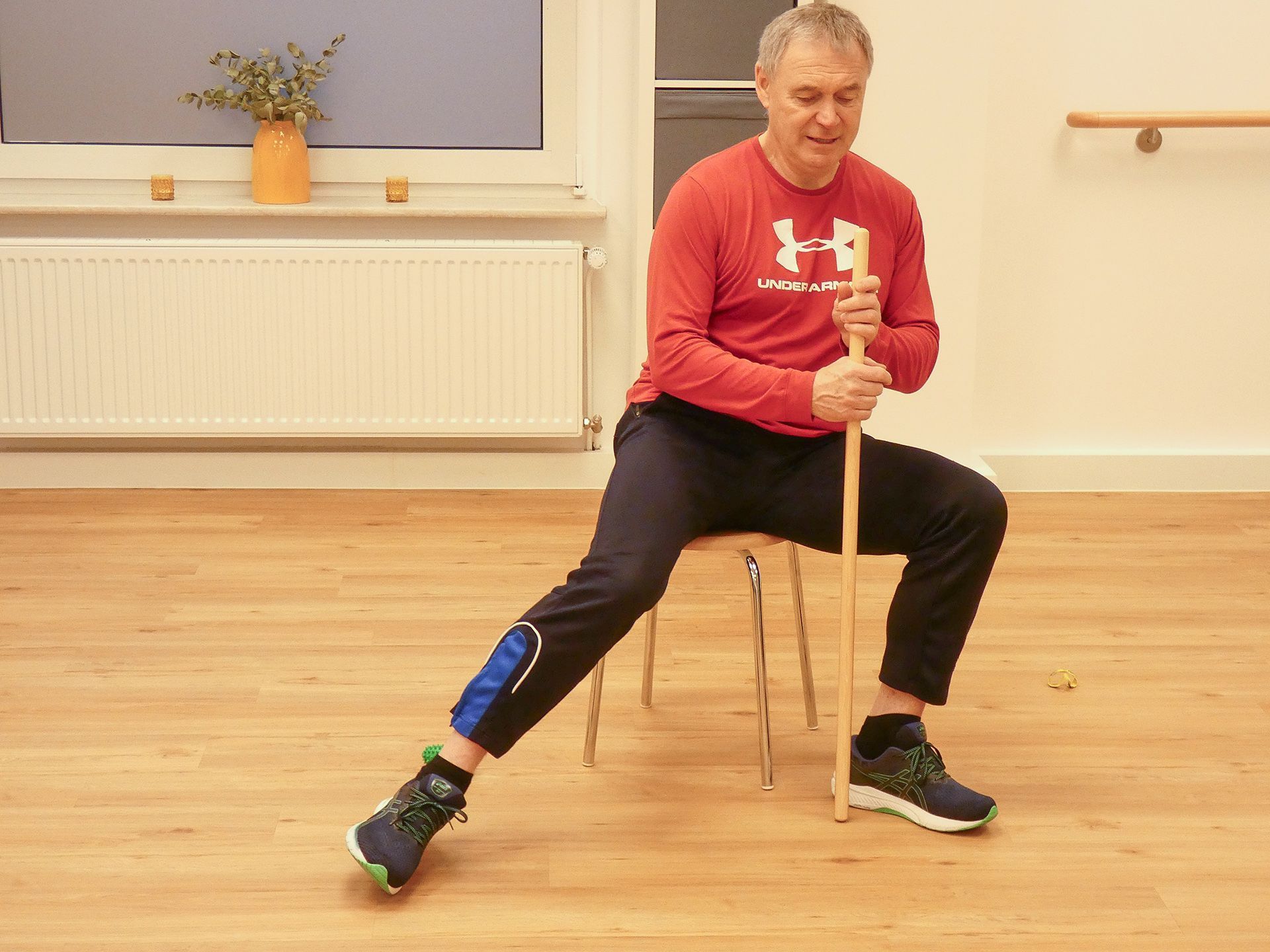 Hockergymnastik im Seminar- und Bewegungsraum der Reha-Sportgruppe Wardenburg. Der Übungsleiter zeigt ein Übung mit Stab.