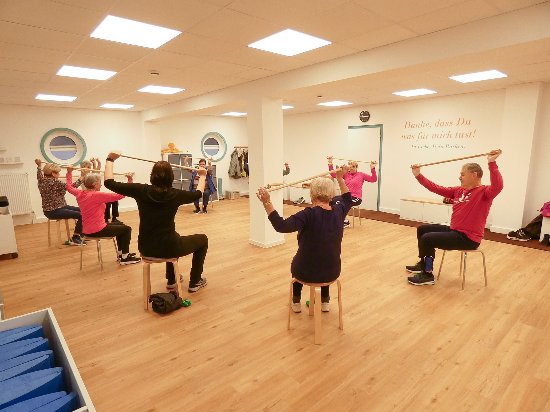 Hockergymnastik im Seminar- und Bewegungsraum der Reha-Sportgruppe Wardenburg. Die Teilnehmer machen eine Übung mit dem Stab.