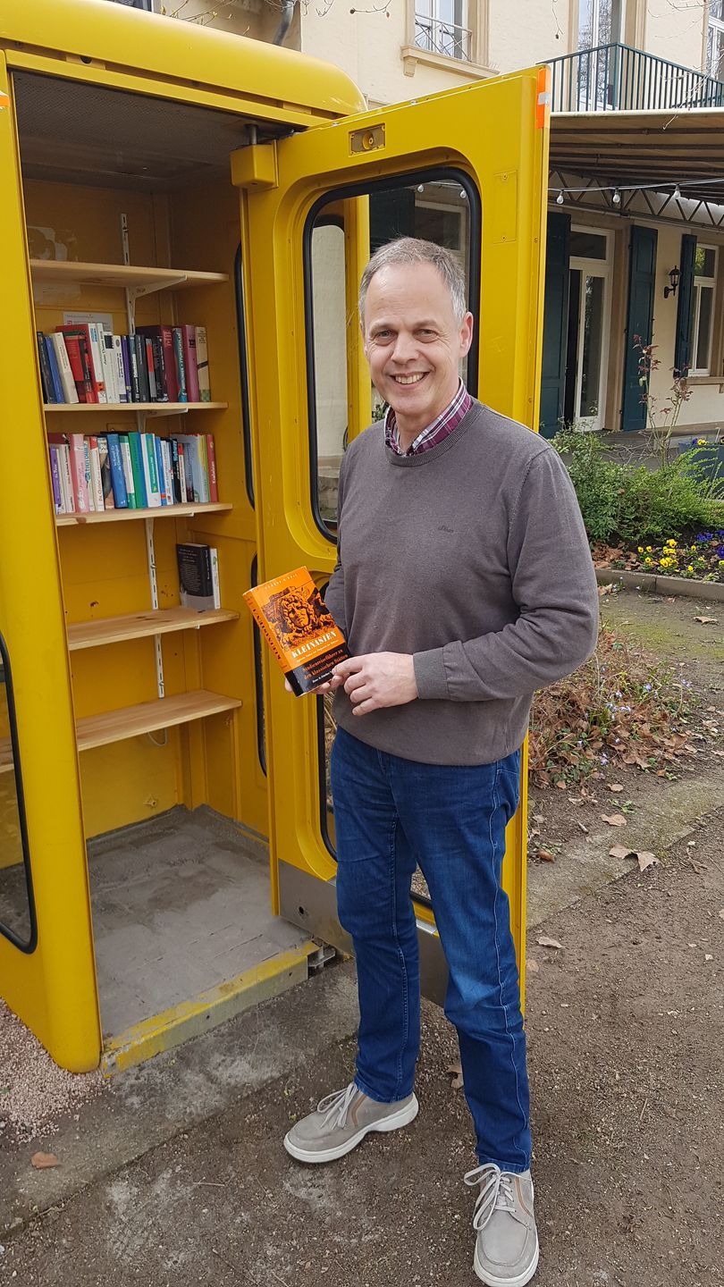 Bürgermeister Jochen Schmitt vor einer gelben Telefonzelle. Die Türe ist offen, man sieht ein halb gefülltes Bücherregal und Herr Schmitt präsentiert ein Buch. Bürgermeister Jochen Schmitt vor einer gelben Telefonzelle. Die Türe ist offen, man sieht ein halb gefülltes Bücherregal und Herr Schmitt präsentiert ein Buch.