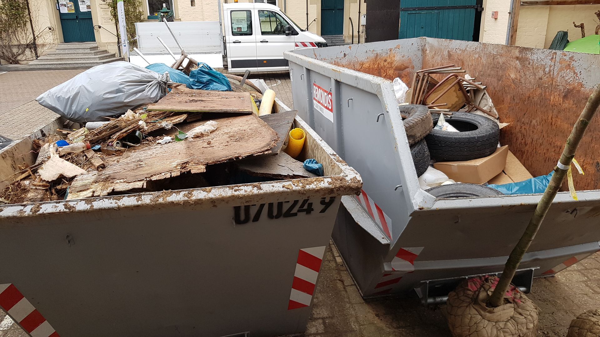 2 große Müllcontainer im Vordergrund gefüllt mit Unrat wie Reifen, Müllsäcke und Möbelresten 2 große Müllcontainer im Vordergrund gefüllt mit Unrat wie Reifen, Müllsäcke und Möbelresten