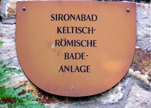 Schild in Wappenfarm auf Natursteinmauer mit der Aufschrift Sirona, Bad keltisch- römische Badeanlage Schild in Wappenfarm auf Natursteinmauer mit der Aufschrift Sirona, Bad keltisch- römische Badeanlage
