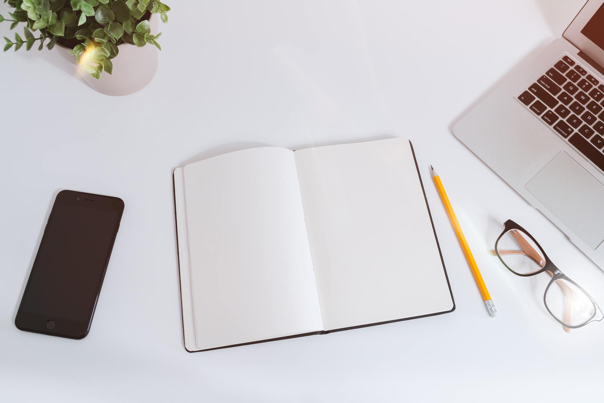 Auf dem Bild steht rechts ein Laptop, darunter liegt eine Brille, daneben liegt ein Stift und ein aufgeschlagenes Notizbuch. Links sieht man noch eine Pflanze und ein Smartphone.
