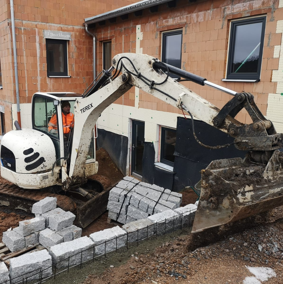 Der Minibagger wird eingesetzt bei z. B. Hinterfüllungen am Bau - auf dem Bild wird eine Mauer gebaut.