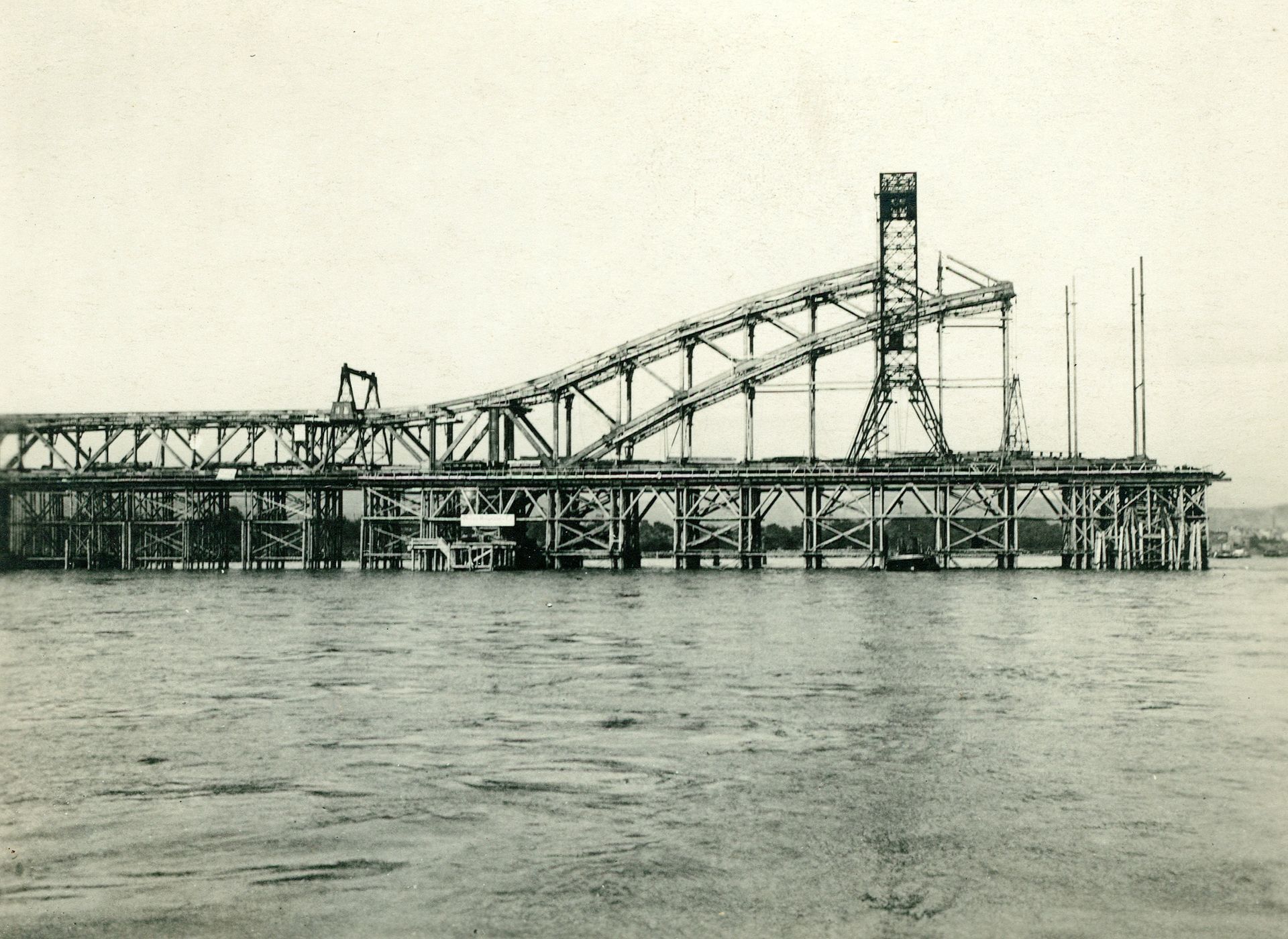 Der Brückenbogenträger entsteht. Kein Teil scheint dem anderen zu gleichen. Die filigran wirkenden Konstruktionselemente werden miteinander vernietet. Auf Holzpfählen, die in den Rheingrund getrieben sind, lagern zunächst die zu montierenden tonnenschweren Eisenteile. Sammlung: Berlin-Brandenburgisches Wirtschaftsarchiv e.V. Der Brückenbogenträger entsteht. Kein Teil scheint dem anderen zu gleichen. Die filigran wirkenden Konstruktionselemente werden miteinander vernietet. Auf Holzpfählen, die in den Rheingrund getrieben sind, lagern zunächst die zu montierenden tonnenschweren Eisenteile.
Sammlung: Berlin-Brandenburgisches Wirtschaftsarchiv e.V.