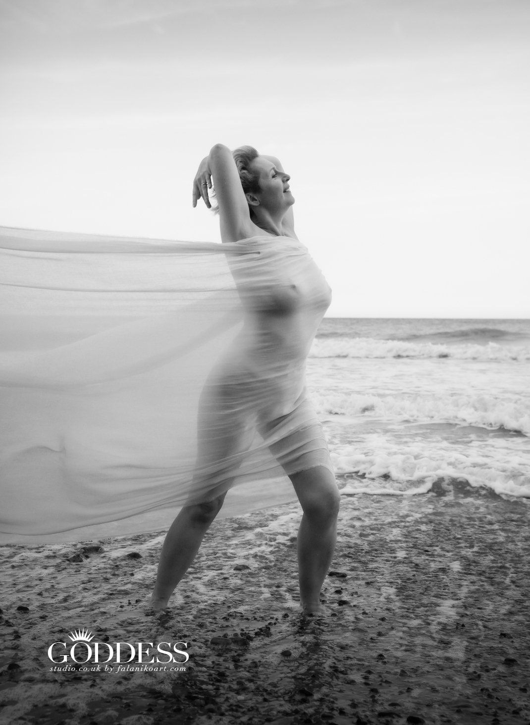 A stunning woman in her sixties stands proudly on a Norfolk beach. She arches back to confidently show her curves, covered by a flowing muslin sheet. Today she celebrates her body.