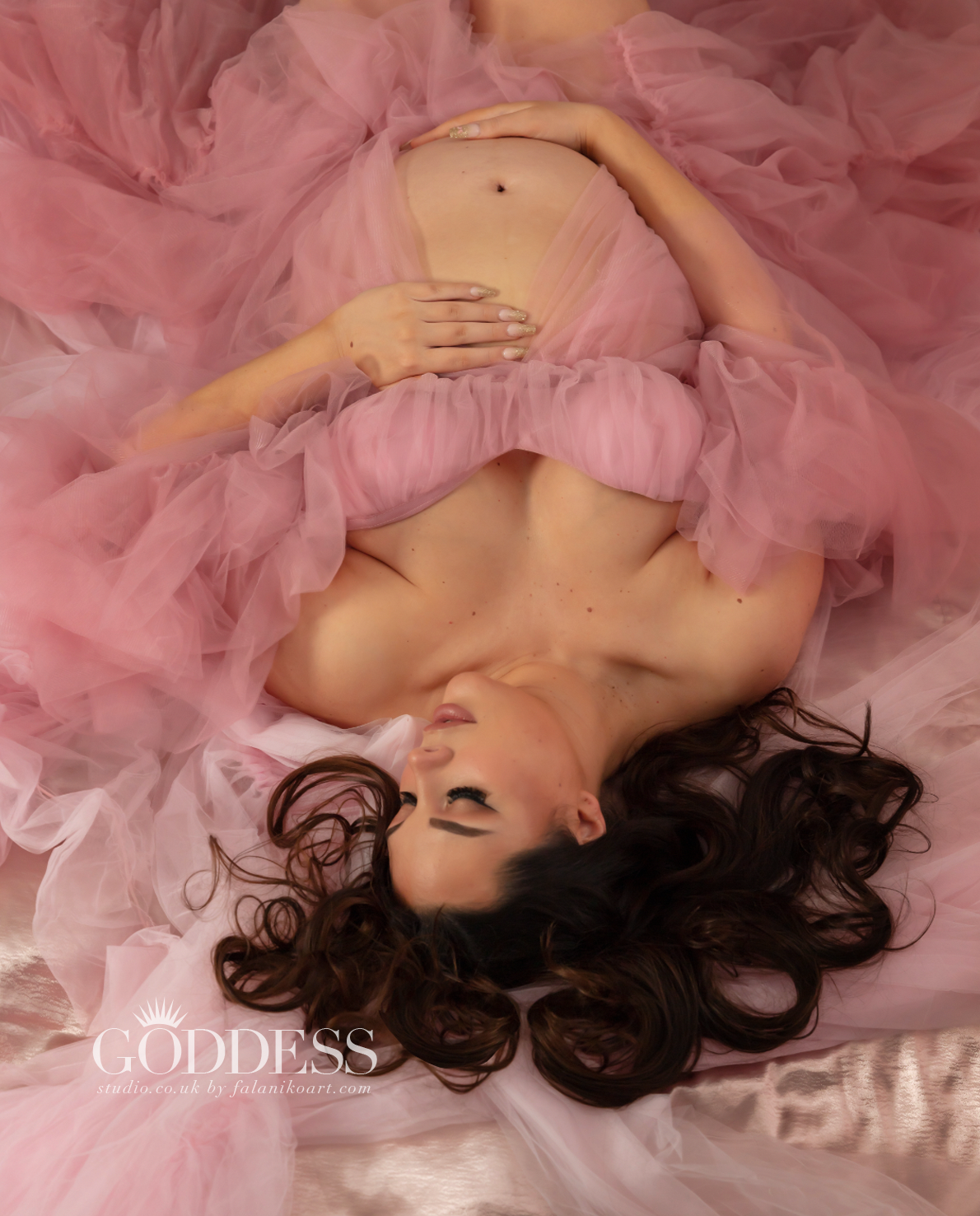 A pregnant woman, dressed in a flowing pink robe, lies on pink satin sheets. Her hair is splayed out and her makeup is flawless. She is fully engaged in her photo session and the special moment of her daughter's coming.