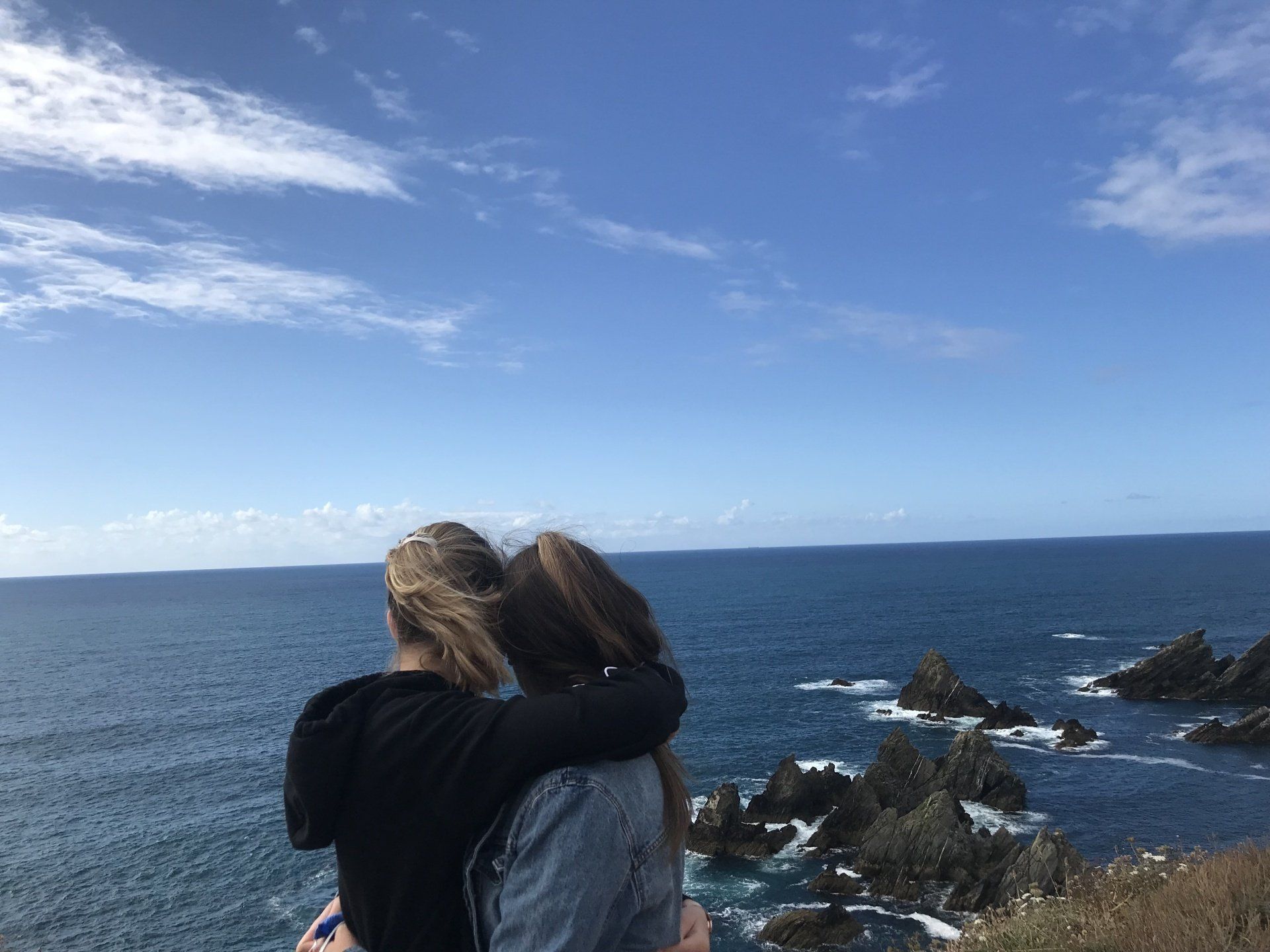 Two girls hugging each other on a cliff as they look at the ocean