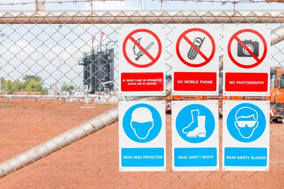 View of industrial site through wire fence. Attached to the fence are 6 showing prohibition and mandatory safety signs, specifically No Naked Lights, No Mobile Phones, No Photography.  Mandatory Hard Hats, Boots and safety Glasses