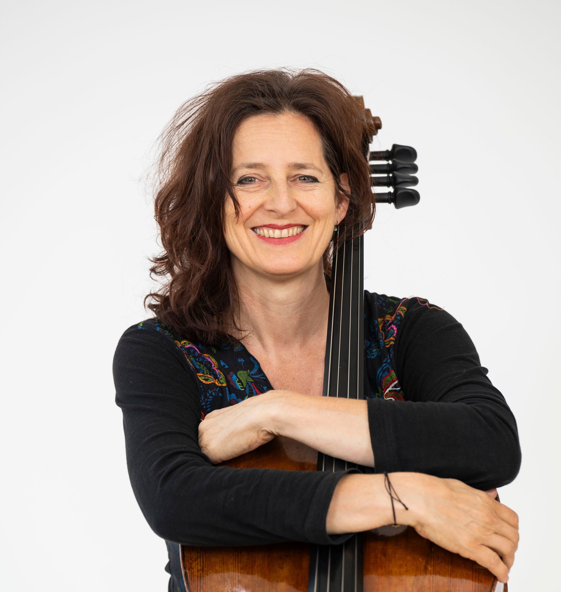 Sue Schlotte sitzt mit locker umarmtem Cello vor einem hellgrauen Hintergrund und lächelt in die Kamera – persönliche Darstellung der Musikerin.