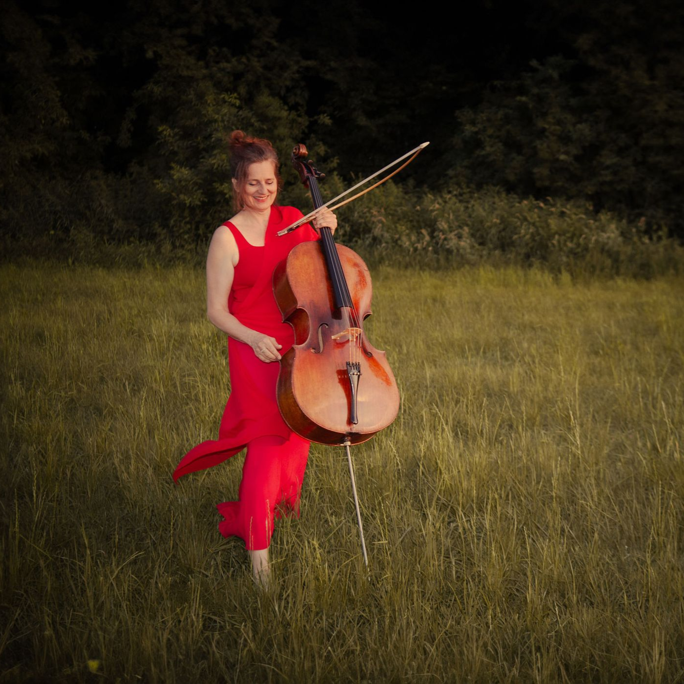 Sue Schlotte, Cellistin und Gründerin des CelloByHeart-Projekts, geht mit Cello in der Hand über eine Wiese bei Sonnenuntergang – Symbol für Musik und Leidenschaft.