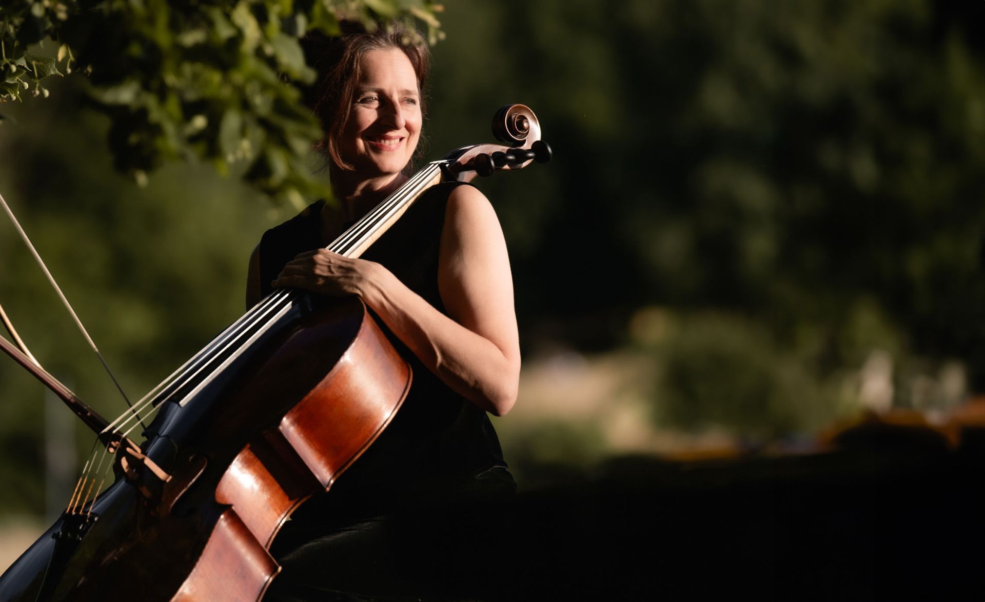Sue Schlotte, Cellistin, beim Cello spielen. Sie steht neben einem Baum, lächelt und verkörpert musikalische Leidenschaft und Ausdruck.