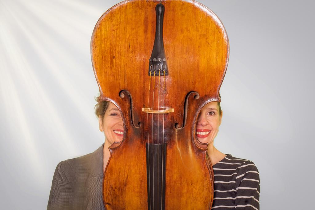 Sue Schlotte und Leana Luzius sitzen vor hellgrauem Hintergrund, getrennt durch ein auf den Kopf gestelltes Cello – symbolträchtiges Porträt des Duos.