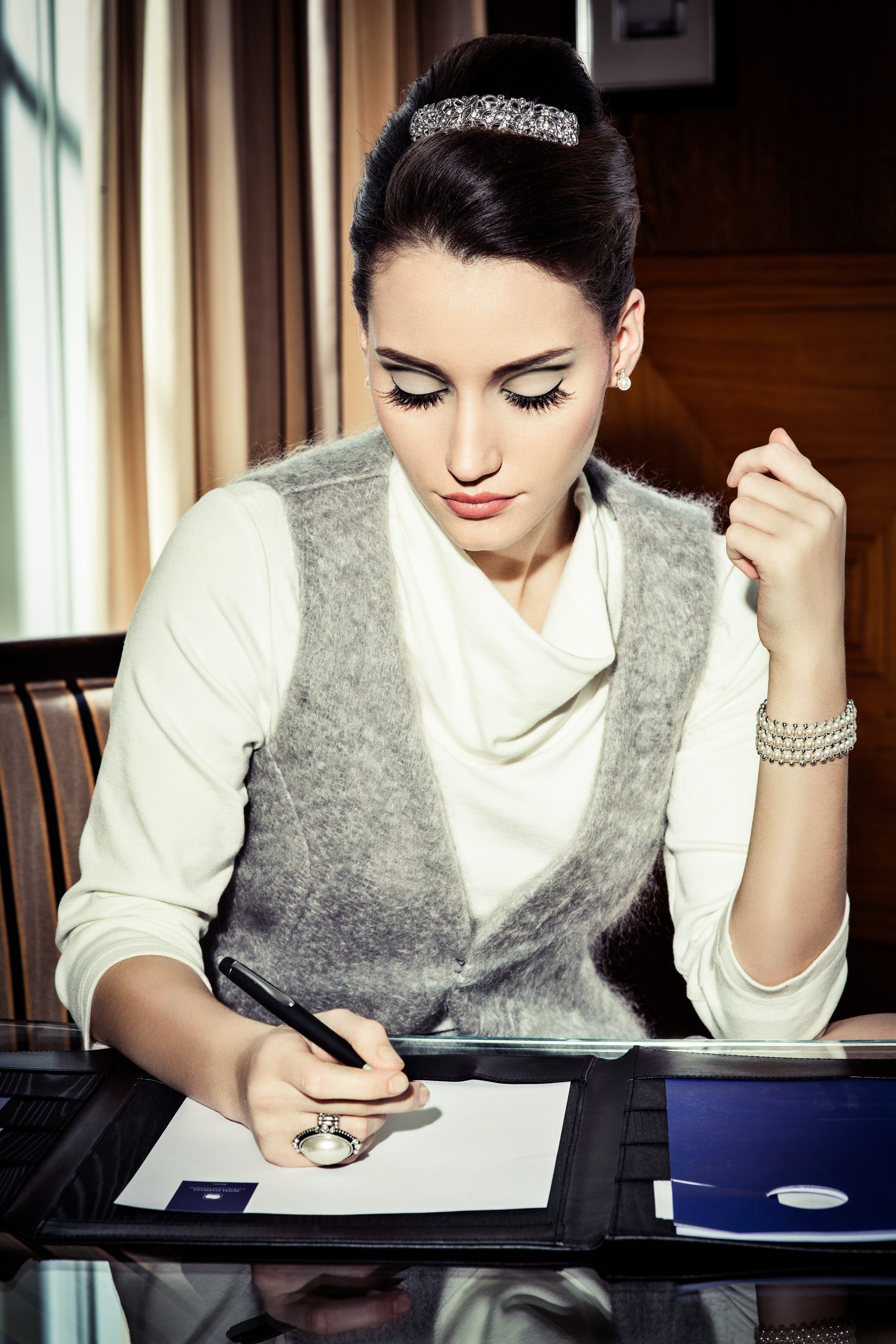 Frau sitzt am Schreibtisch und schreibt mit einem schwarzen Stift auf ein weißes Blatt Papier. Sie trägt ein weißes Oberteil mit einer grauen Weste darüber. Marke der Kleidung ist 3 Zehn.