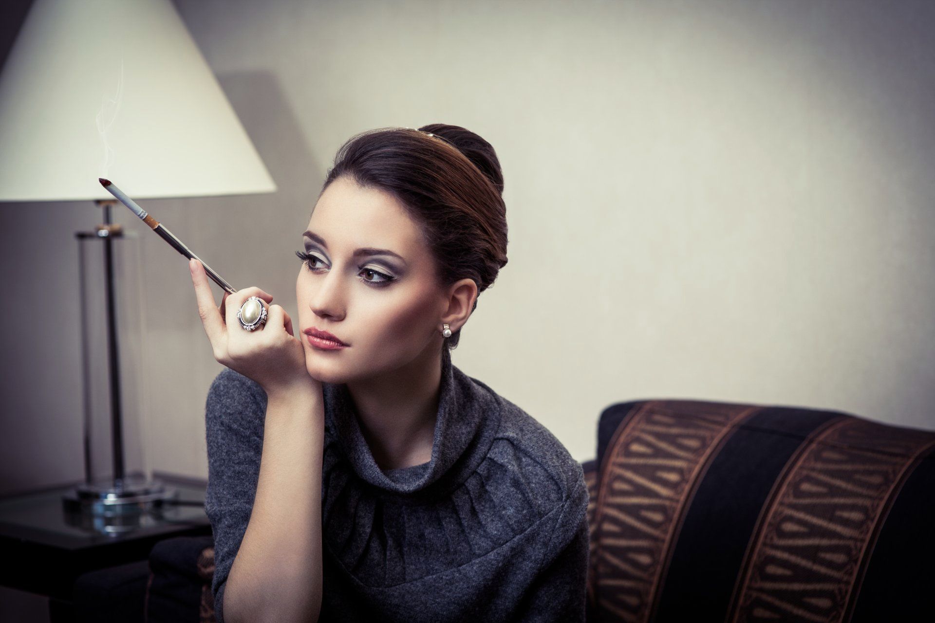 Frau sitzt auf Hocker und hält eine Zigarettenspitze in der rechten Hand. Marke des grauen Pullovers ist 3 Zehn. Gala Makeup