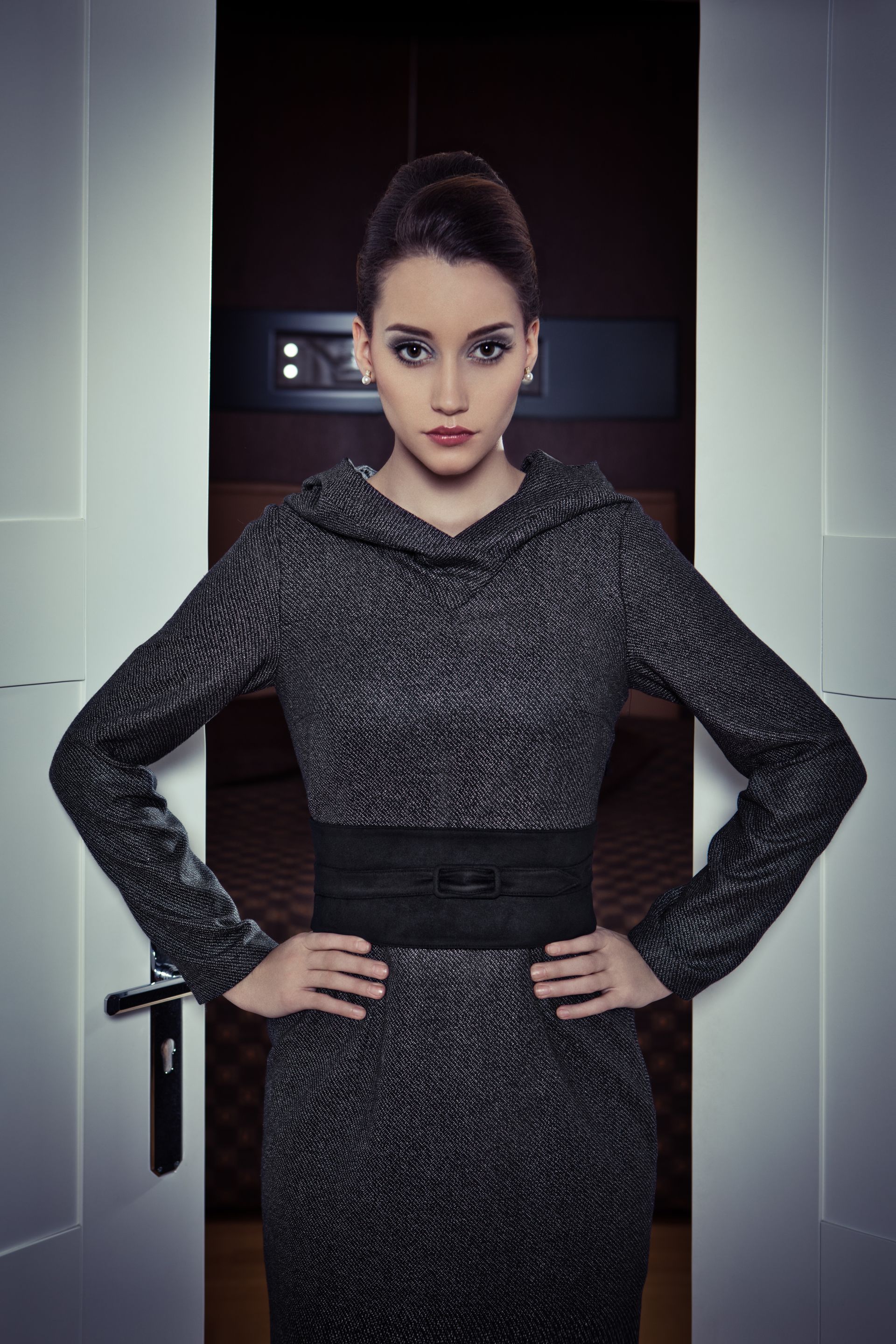 Frau steht vor einer Tür und trägt ein graues Kleid mit schwarzem Gürtel. Marke der Kleidung ist 3 Zehn.