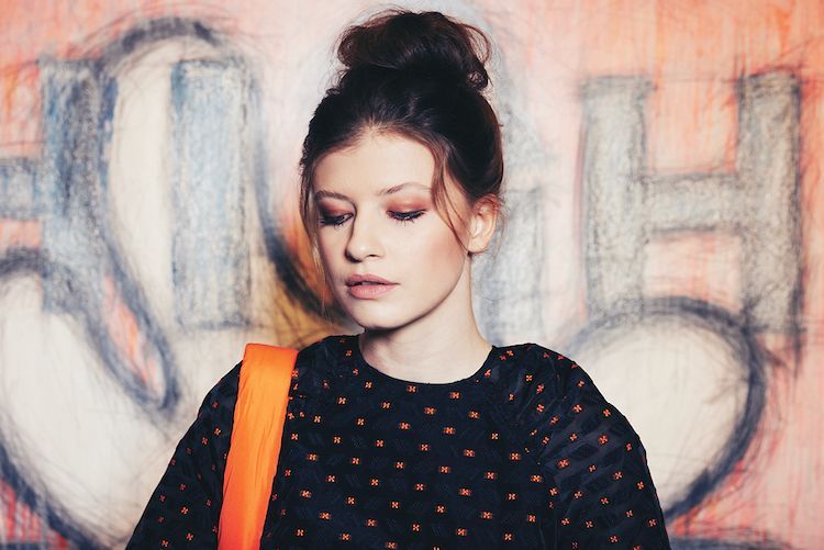 Porträt einer Frau mit hochgestecktem Dutt trägt einen dunkelblauen Pullover der Marke Coster Copenhagen mit orangenen Punkten und eine orangefarbene Handtasche.