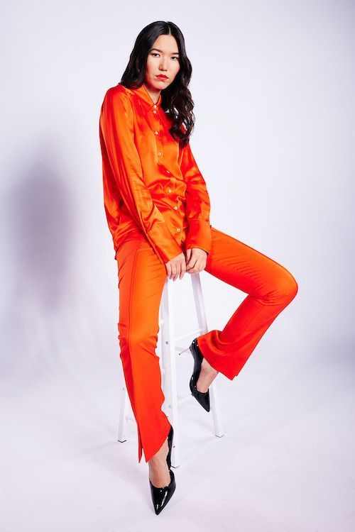 Frau mit schwarzen Haaren und rotem Lippenstift trägt einen orangefarbenen Anzug der Marke Patrizia Pepeund sitzt auf einem Barhocker vor weißem Hintergrund.