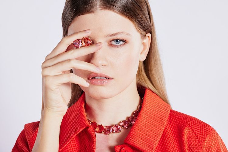 Frau mit langen Haaren und roter Bluse trägt eine Kette mit roten Steinen und hält sich ein Schmuckstück vor ihr rechtes Auge. Marke: Swarovski