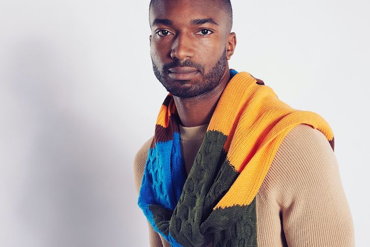Fascionshooting mit Männermodel mit sehr kurzen Haaren und dunkler Haut, trägt einen beigen Pullover und einen bunten Schal der Marke Zara.