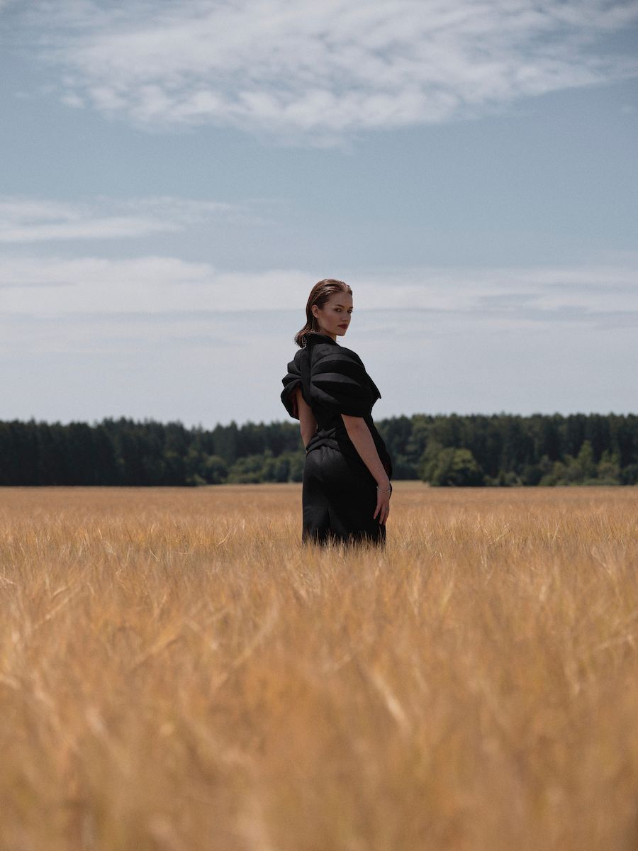 Eine junge Dame mit langen, nach hinten gestylten Haaren steht im Kornfeld. Sie trägt ein schwarzes Kleid.