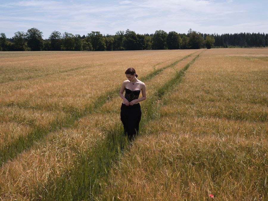 Eine junge Dame steht in einem Kornfeld und blickt auf den Boden. Sie trägt einen Pferdeschwanz und ein schwarzes, trägerloses Oberteil und einen schwarzen, engen Rock.
